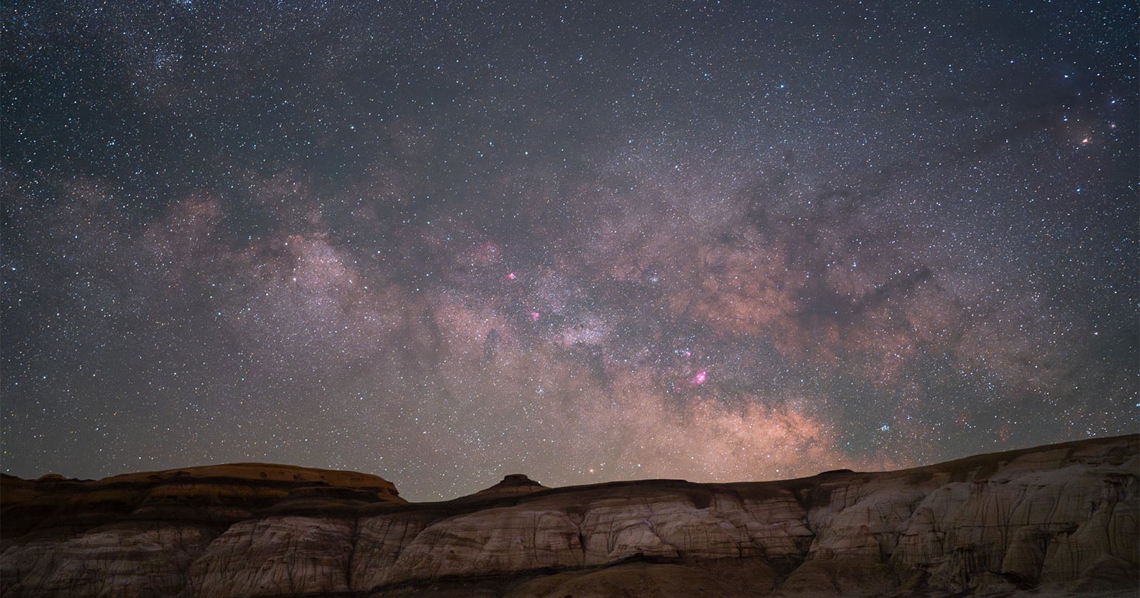 A star-filled night sky with the Milky Way galaxy glowing above rugged, rocky cliffs in the foreground. The scene highlights bright stars, cosmic dust, and soft, colorful nebulae stretching across the horizon.