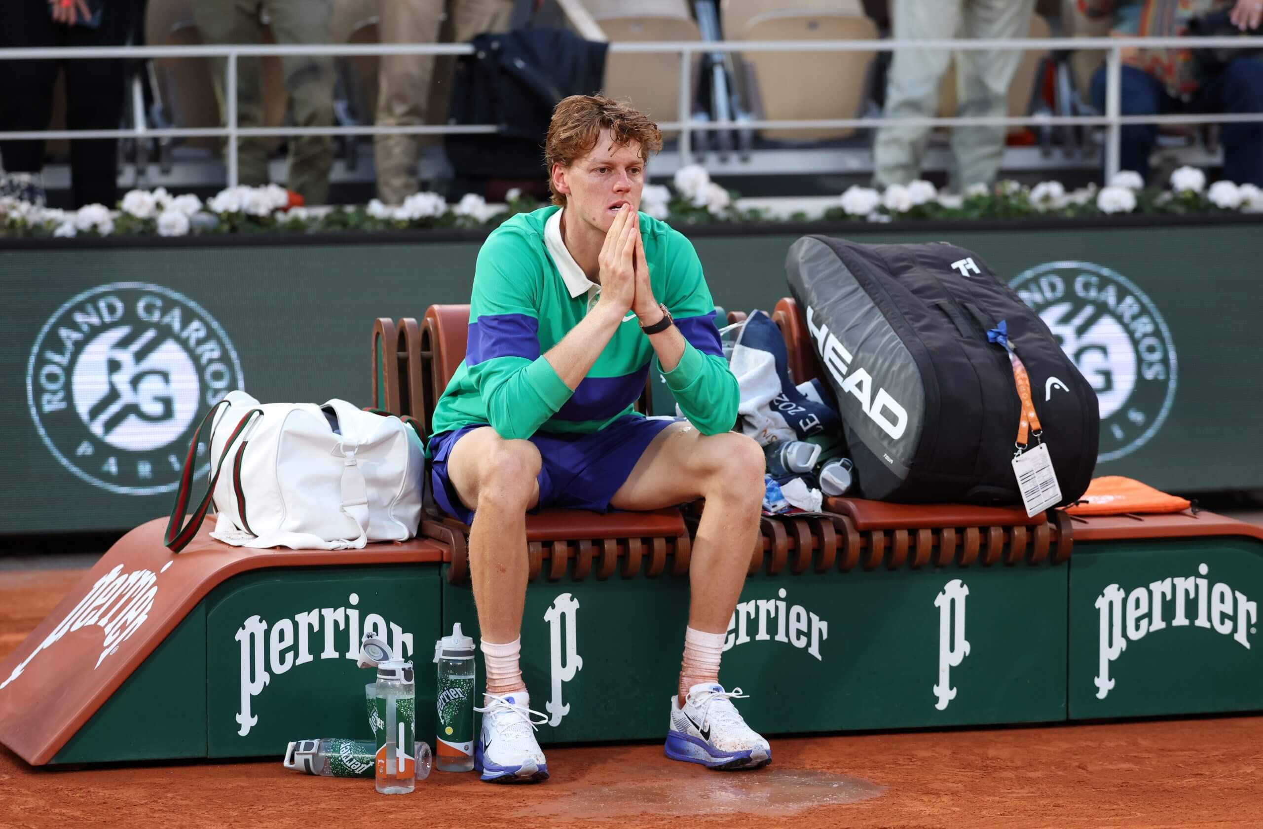 Jannik Sinner sits disconsolately on a bench wearing a green-and-blue tennis outfit.