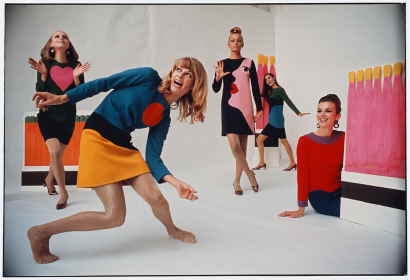 Four women in colorful, graphic mod dresses pose playfully in a white room with large, bright matchstick props in the background. Their poses and expressions are energetic and dynamic, evoking a 1960s fashion vibe.