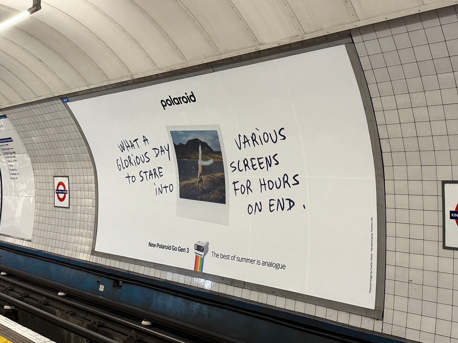 A Polaroid ad in a subway station shows a photo of a mountain landscape with handwritten text: “What a glorious day to stare into various screens for hours on end.” London Underground signs are visible on the tiled wall.