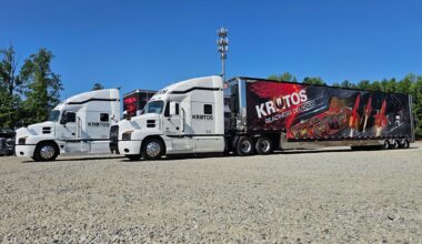 Line of white semi trucks parked in a row on a gravel lot beneath a clear blue sky, with a large Kratos trailer featuring red and black graphics behind them.