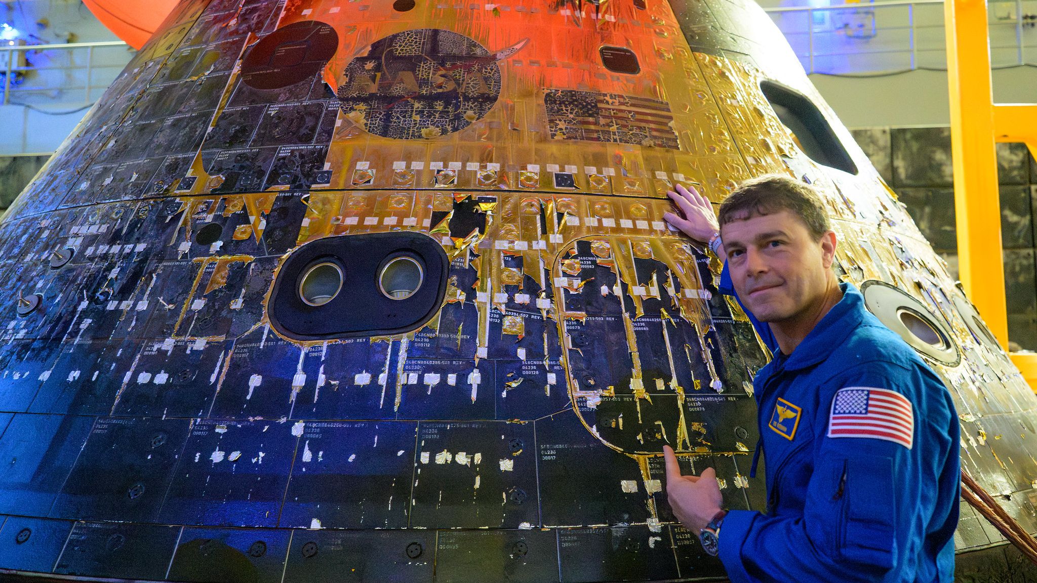 Artemis 2 commander Reid Wiseman is photographed in a blue NASA jumpsuit looking back at the camera with his hand on the charred Orion spacecraft as it sits in the well deck of a recovery ship.