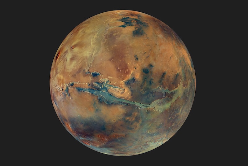 Reddish planet with darker regions on its rocky surface. A long canyon cuts across just south of the equator.