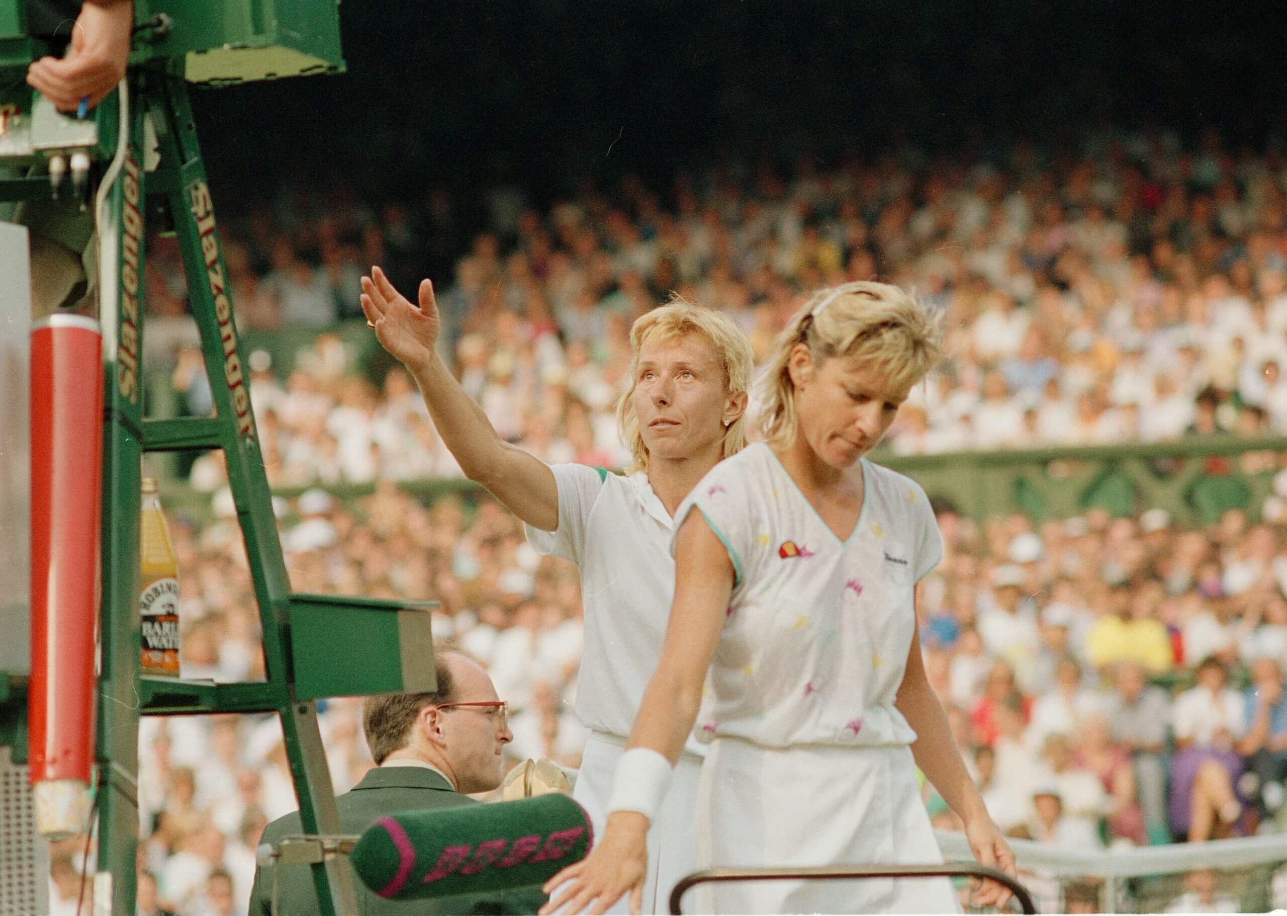 Martina Navratilova (left) raises her right hand to shake hands with an umpire (out of shot) on a green chair, with Chris Evert walking to her left just in front.