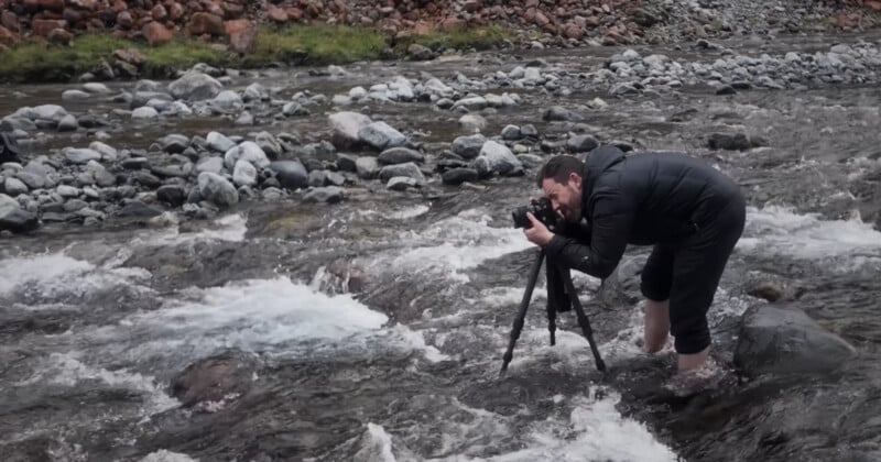 A person in a black jacket stands barefoot in a rocky stream, adjusting a camera on a tripod to take a photo. The water flows around their legs, with rocks and grass visible on the riverbank.