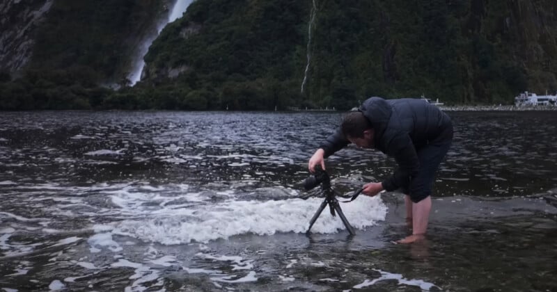 A person stands barefoot in shallow water, adjusting a camera on a tripod as waves approach. Behind them are tree-covered mountains and a waterfall. The sky is overcast, creating a moody, natural atmosphere.