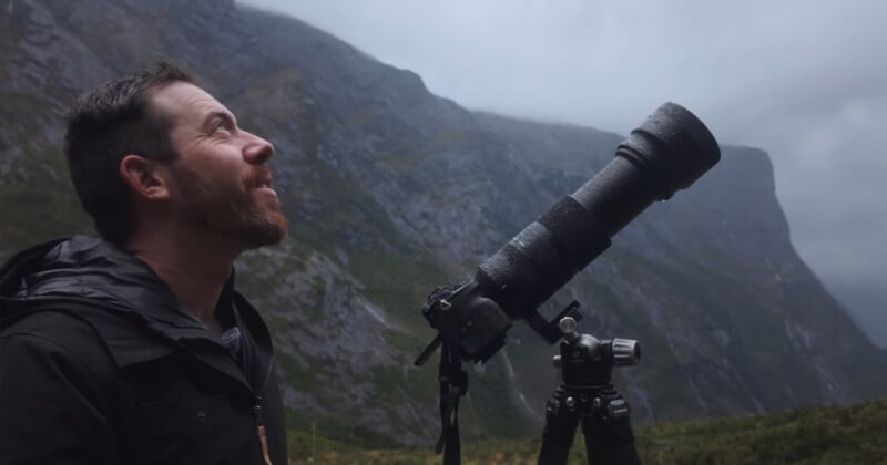 A man with a beard stands outdoors beside a large camera with a telephoto lens on a tripod, looking up at misty mountains in the background.