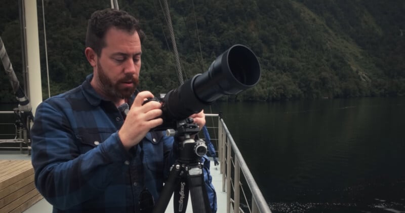A man wearing a blue plaid shirt adjusts a large camera with a telephoto lens on a tripod, standing on the deck of a boat with forested hills and calm water in the background.