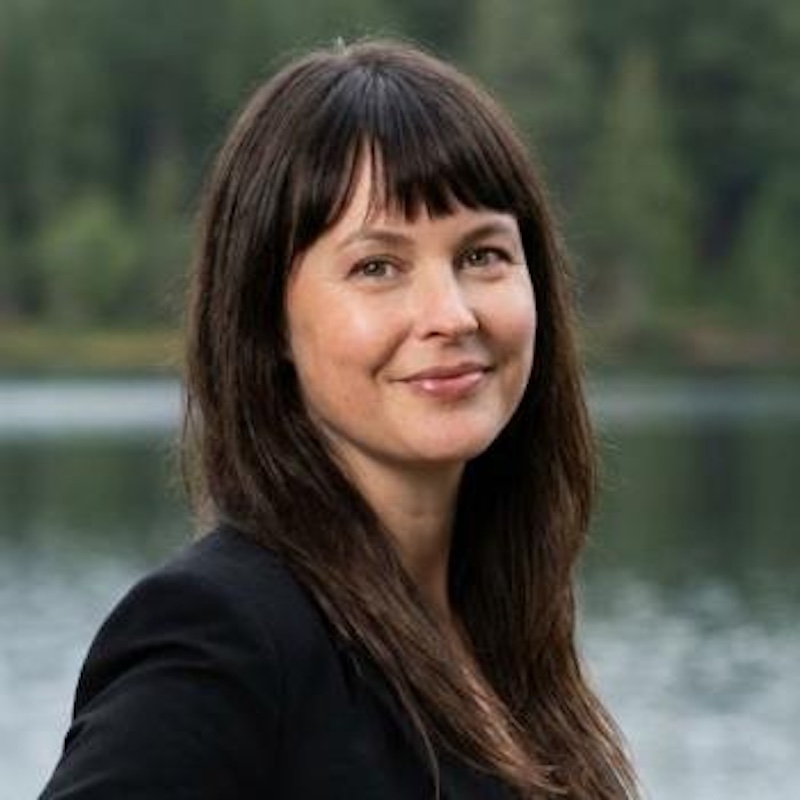 Smiling young woman with long dark hair and bangs and a lake behind her.