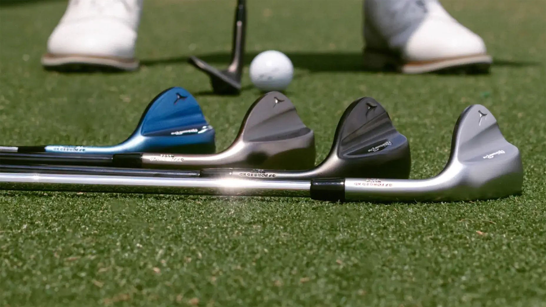 Four golf clubs with different colored heads (blue, black, bronze, and silver) are lined up on grass near a golf ball and tee, with a golfers shoes visible in the background.