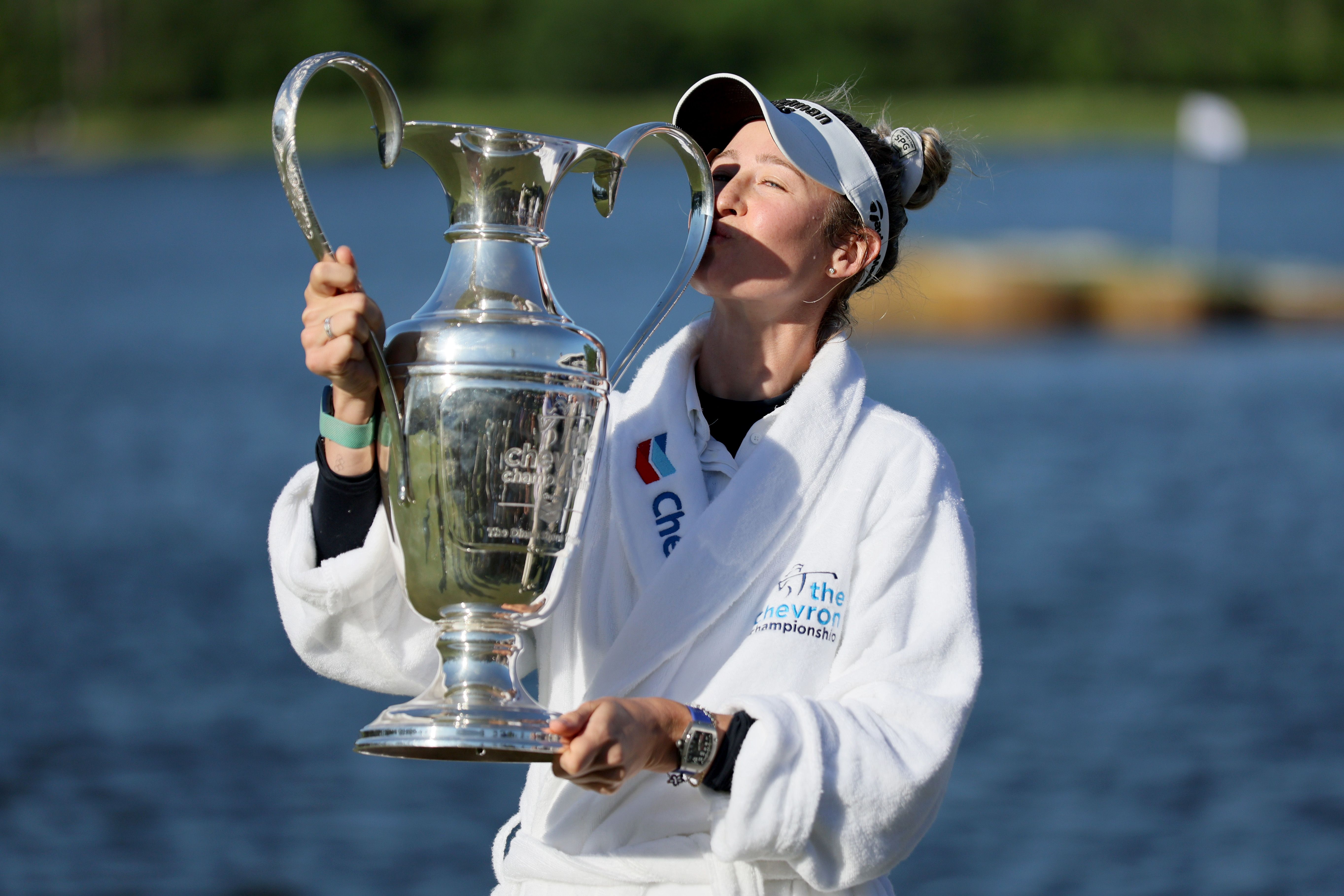 Athlete in white robe triumphantly holds large silver trophy by water, looking up.