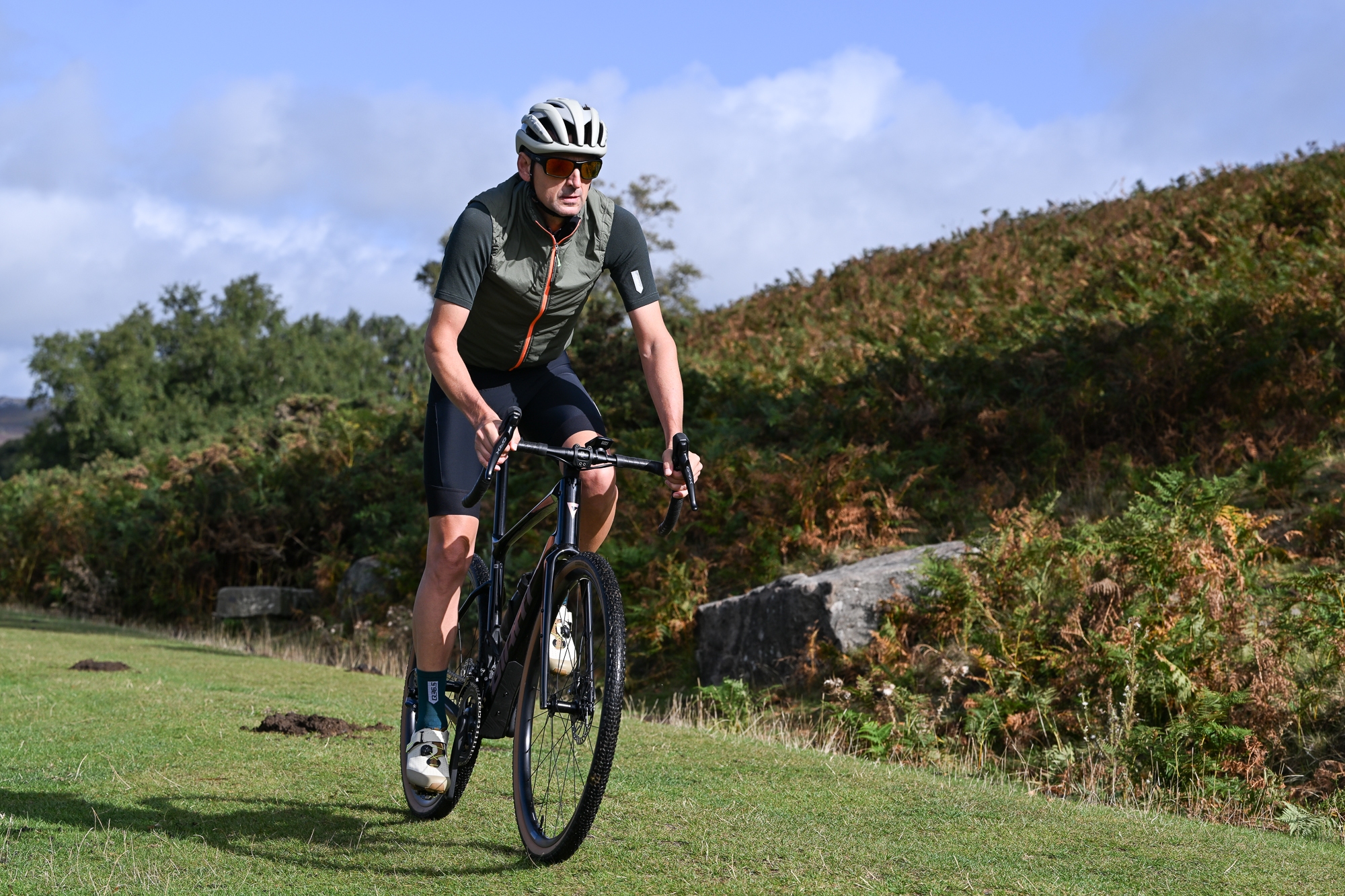 Man wearing black shorts, green jersey and a green gilet riding towards the camera on a black bike on a grassy trail