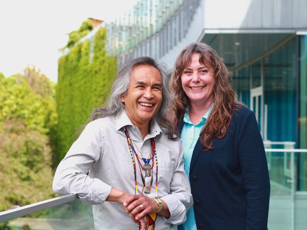 Two people stand in front of an ivy-covered building. One person has long white hair and medium skin tone. The other person has long brown hair and light skin tone.