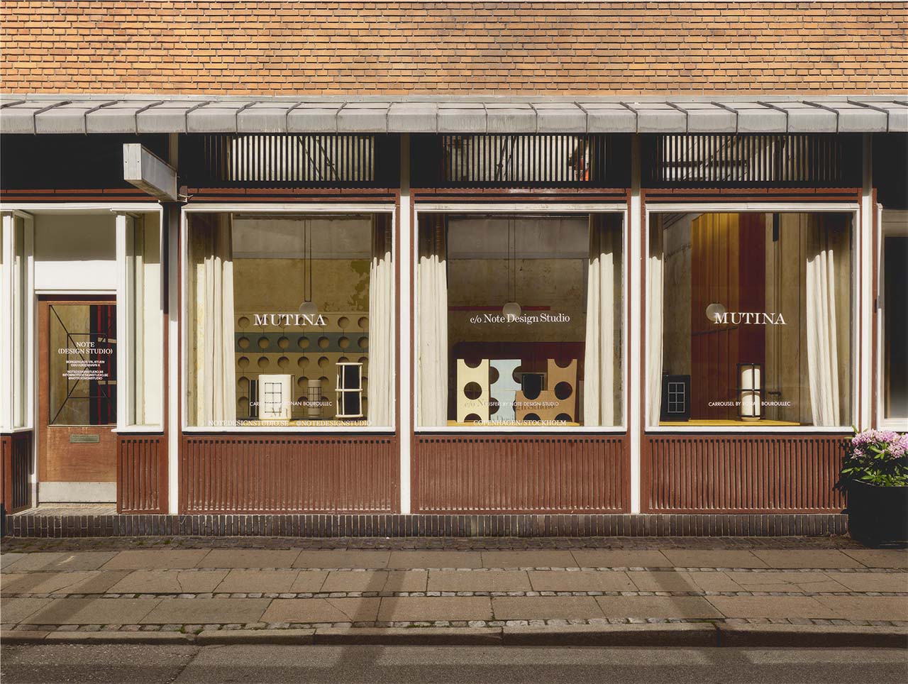 Street view of a storefront with large windows displaying "MUTINA" and "66 New Design Studio" signage, brown and cream exterior, and a potted plant on the right.