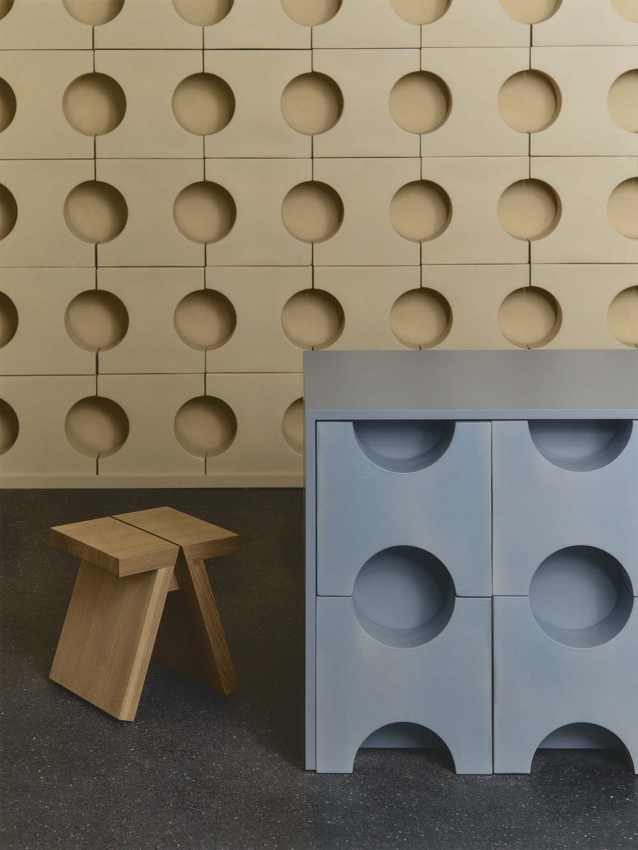 A beige wall with a circular cutout pattern, a light blue cabinet with similar circular designs, and a small wooden stool on a dark floor.