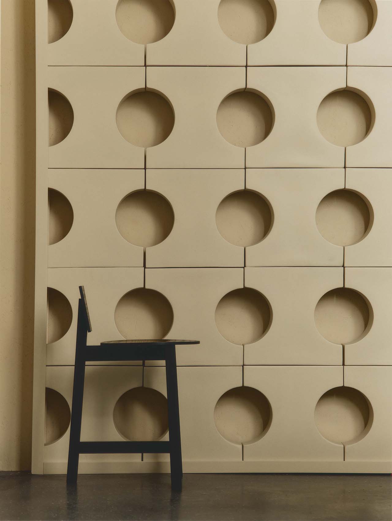 A black chair is positioned in front of a beige wall with a geometric pattern of large circular indents.