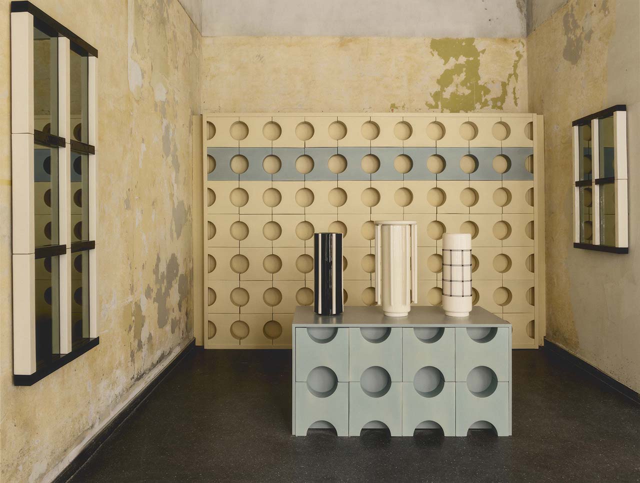 A room with textured yellow walls, featuring modular furniture with circular cutouts, a blue table, three cylindrical vases, and two large wall mirrors.