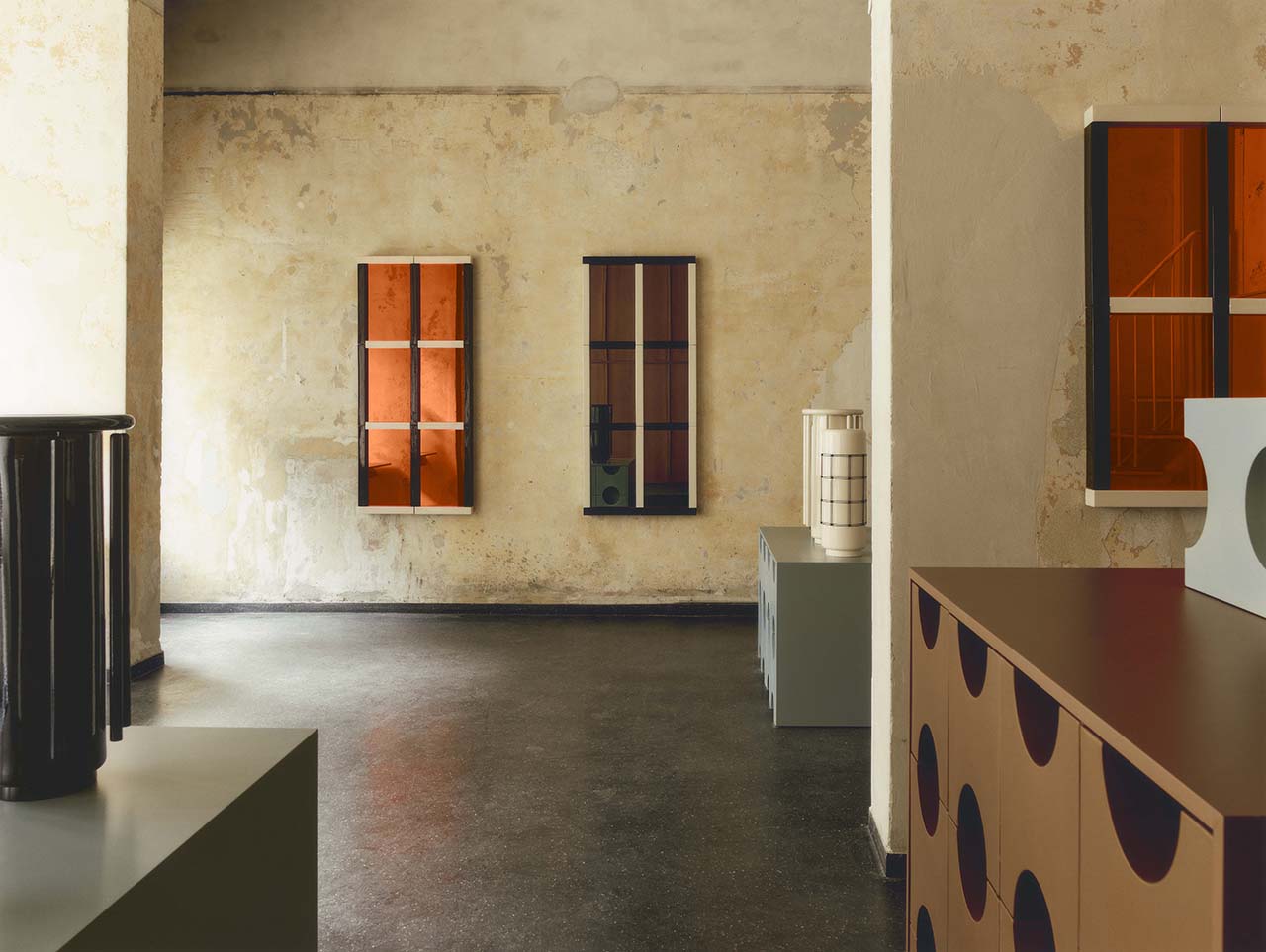 Minimalist interior with textured beige walls, dark floor, geometric furniture, and three rectangular wall-mounted shelves with orange accents.