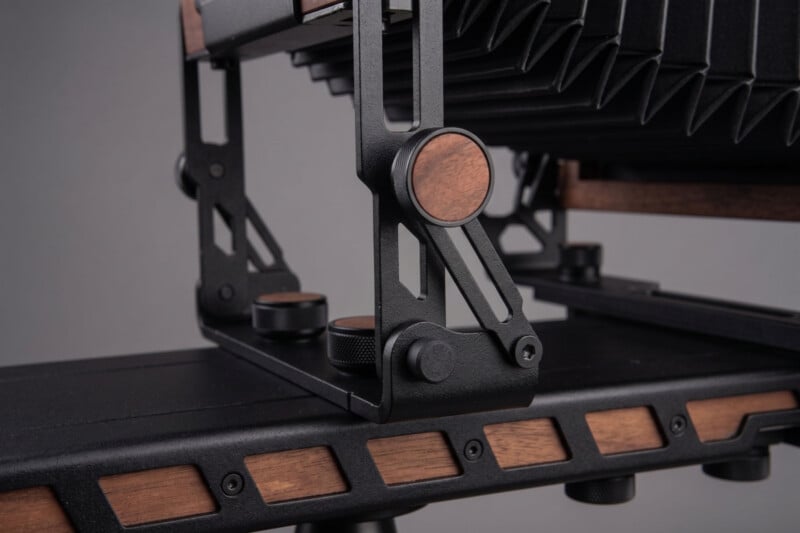 Close-up of a large format camera with black metal and wooden accents, showing detailed knobs, gears, and a bellows mechanism on a smooth, dark base.