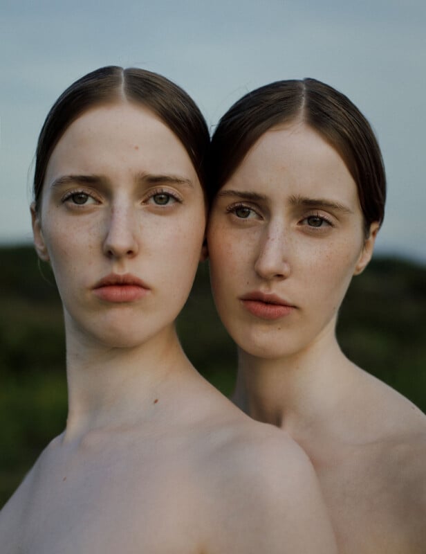 Two young women with similar features, pale skin, and straight brown hair parted in the middle, stand closely together outdoors with a blurred green background. Both have neutral expressions and bare shoulders.