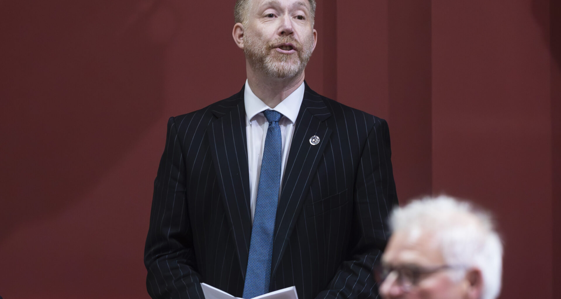 Youri Chassin stands and speaks with papers in his hands at a legislature desk.