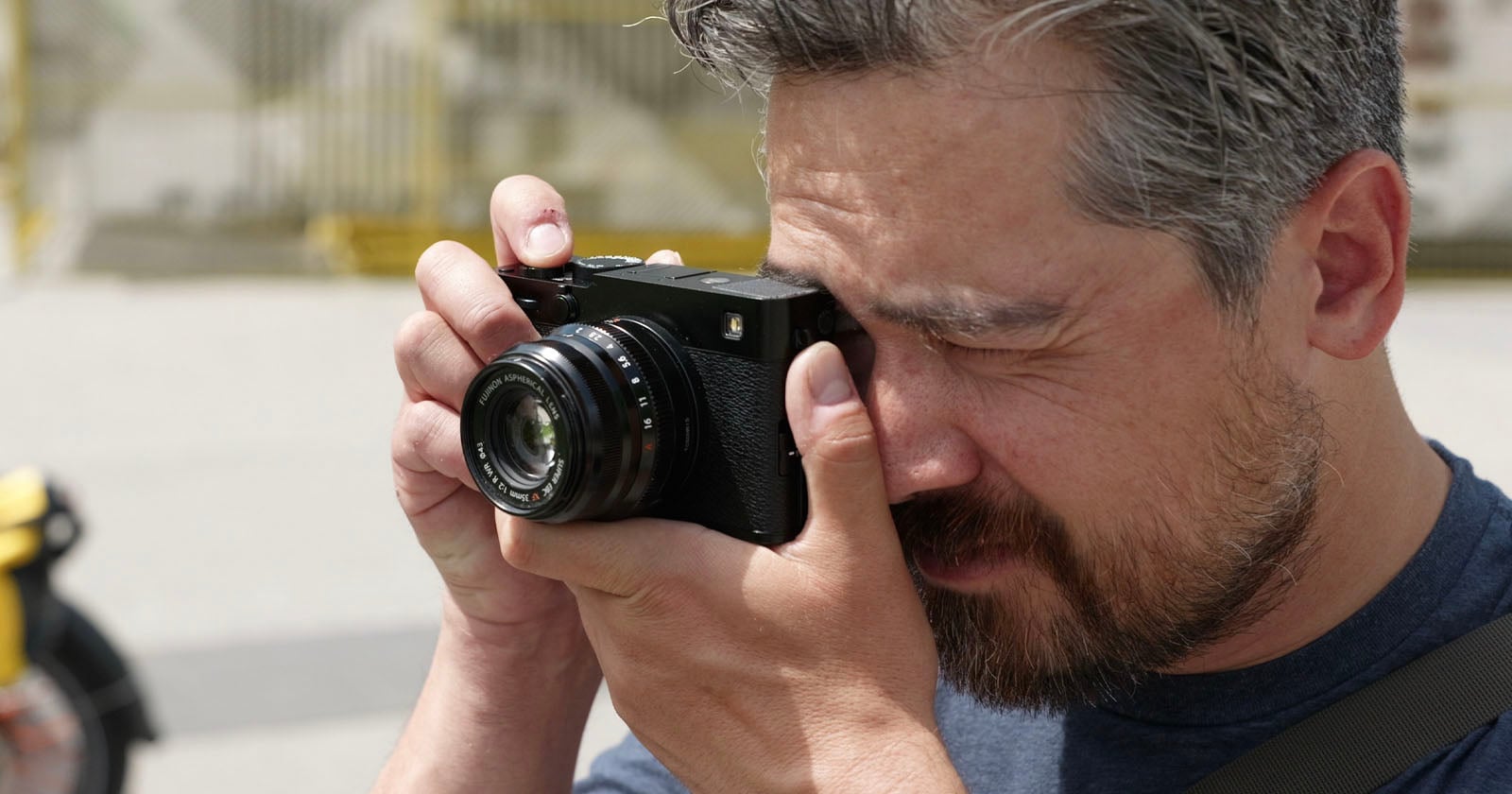 A man with short hair and a beard is holding a black camera up to his face, looking through the viewfinder as he takes a photo outdoors in bright daylight.