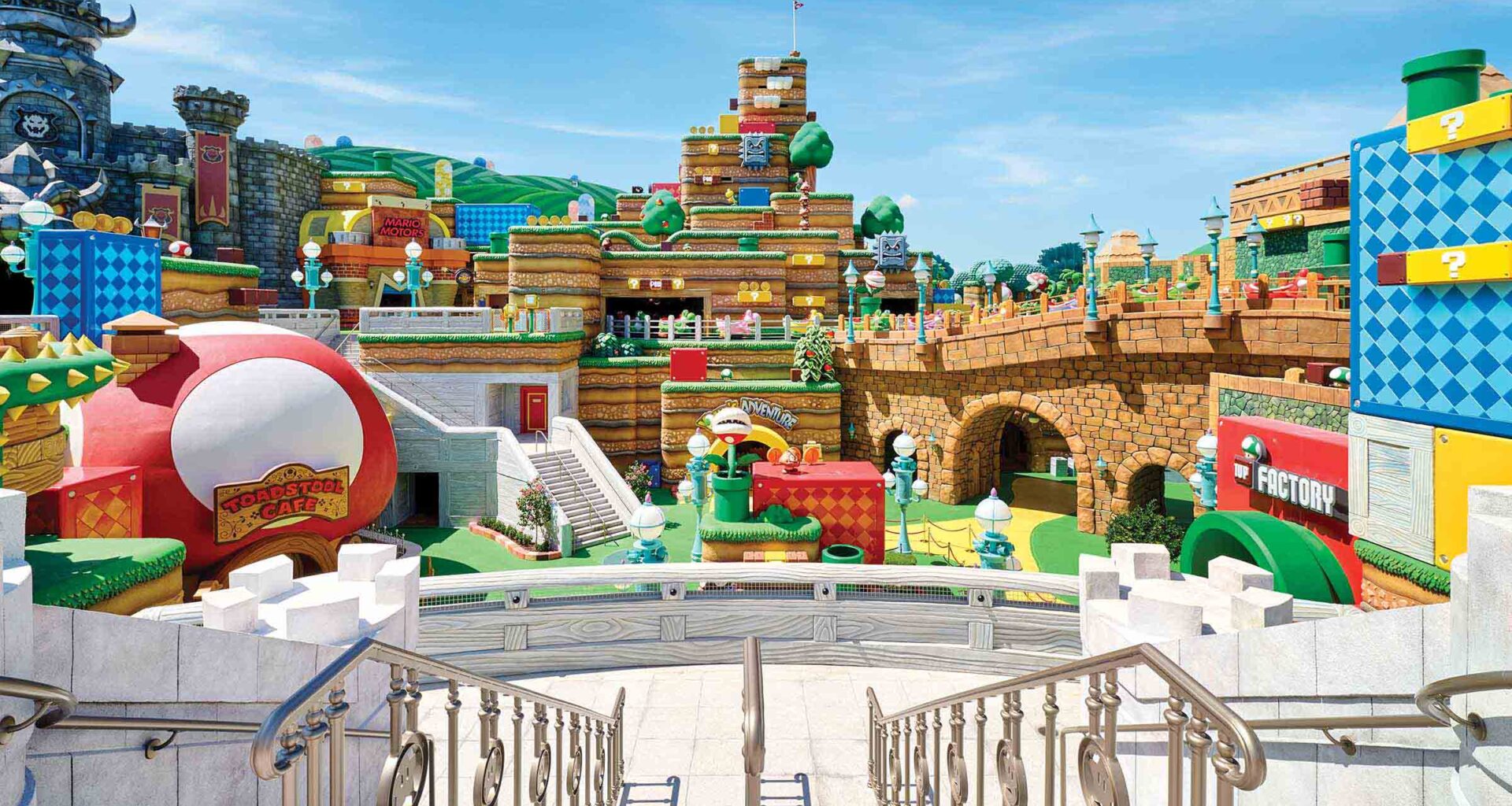 The image shows a section of Super Nintendo World at Universal Orlando Resort. A multilevel landscape is visible, full of Super Mario iconography, including a castle, pipes, platforms, coins, blocks, Thwomps, Goombas, and Piranha Plants, all in the traditional bright colors of Super Mario games. In the lower left of the image, the Toadstool Café has a round, red-and-white roof. On the right side of the image, a large stone bridge is lined with lamp posts and features a Yoshi-themed ride. Throughout the landscape, there are a number of doors and walkways that lead to other sections of the experience.