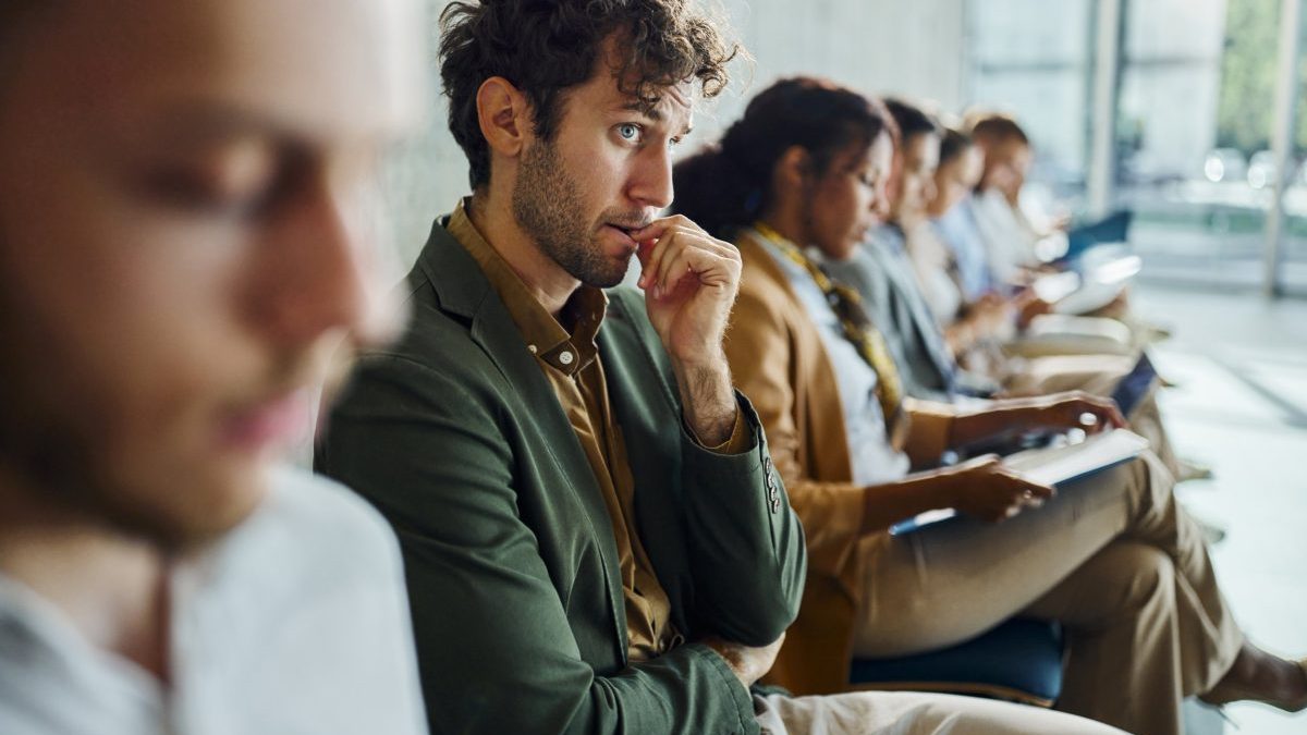 Worried businessman biting his nail while waiting for a job interview with other candidates in a hallway of an office building.