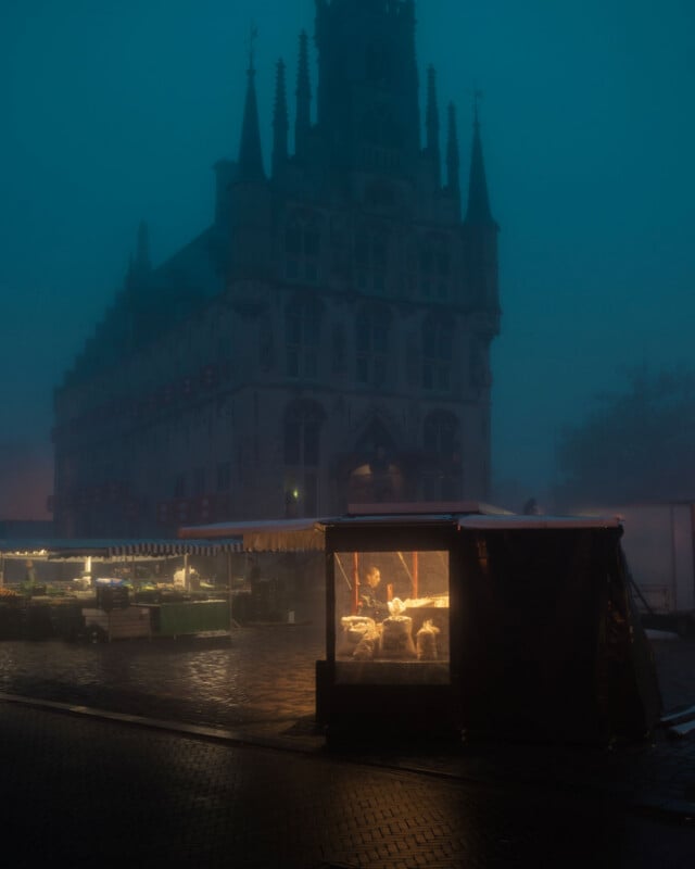 A warmly lit market stall stands on a wet, cobblestone street in front of a tall, gothic-style building shrouded in blue evening fog. Few other stalls are visible in the dim, misty atmosphere.