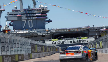 Race car on a track with a large naval aircraft carrier in the background and decorative flags overhead.