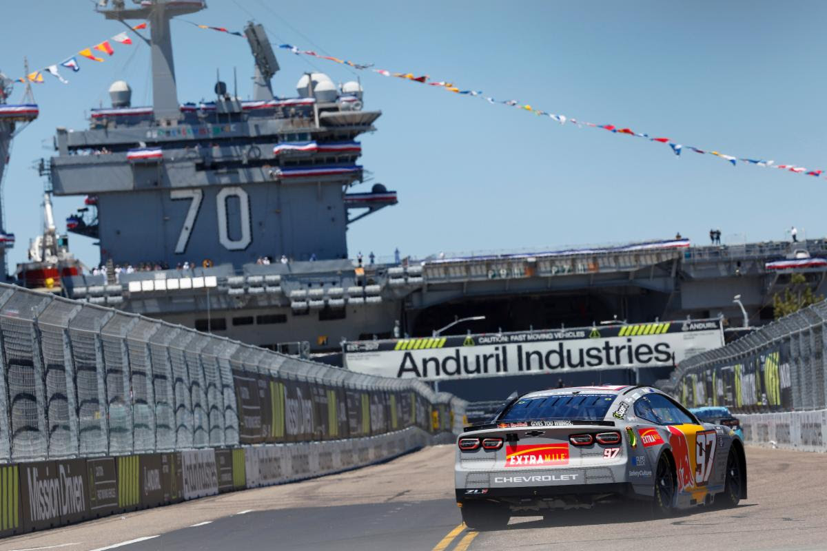 Race car on a track with a large naval aircraft carrier in the background and decorative flags overhead.
