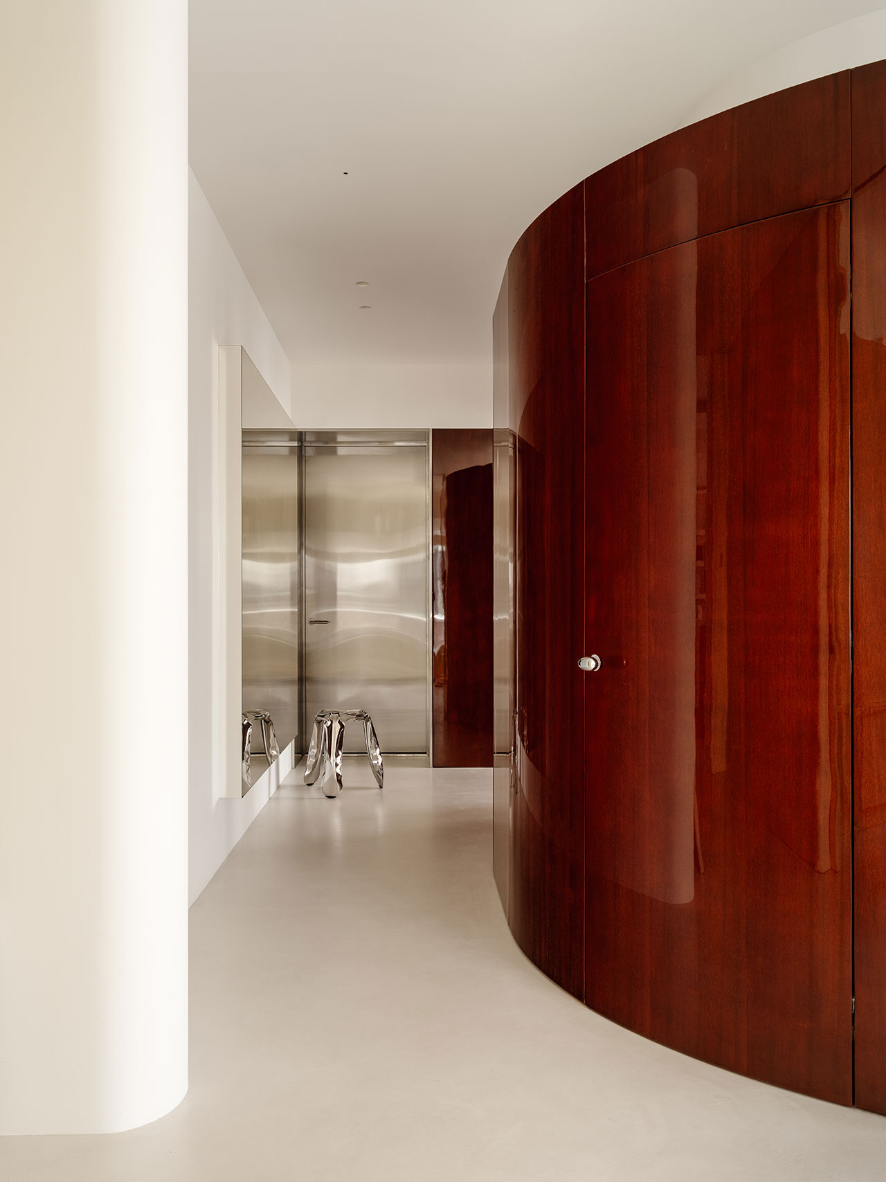 Minimalist hallway with a polished wood curved wall, white floor and walls, a stainless steel door, and an Antwe reflective metallic stool.