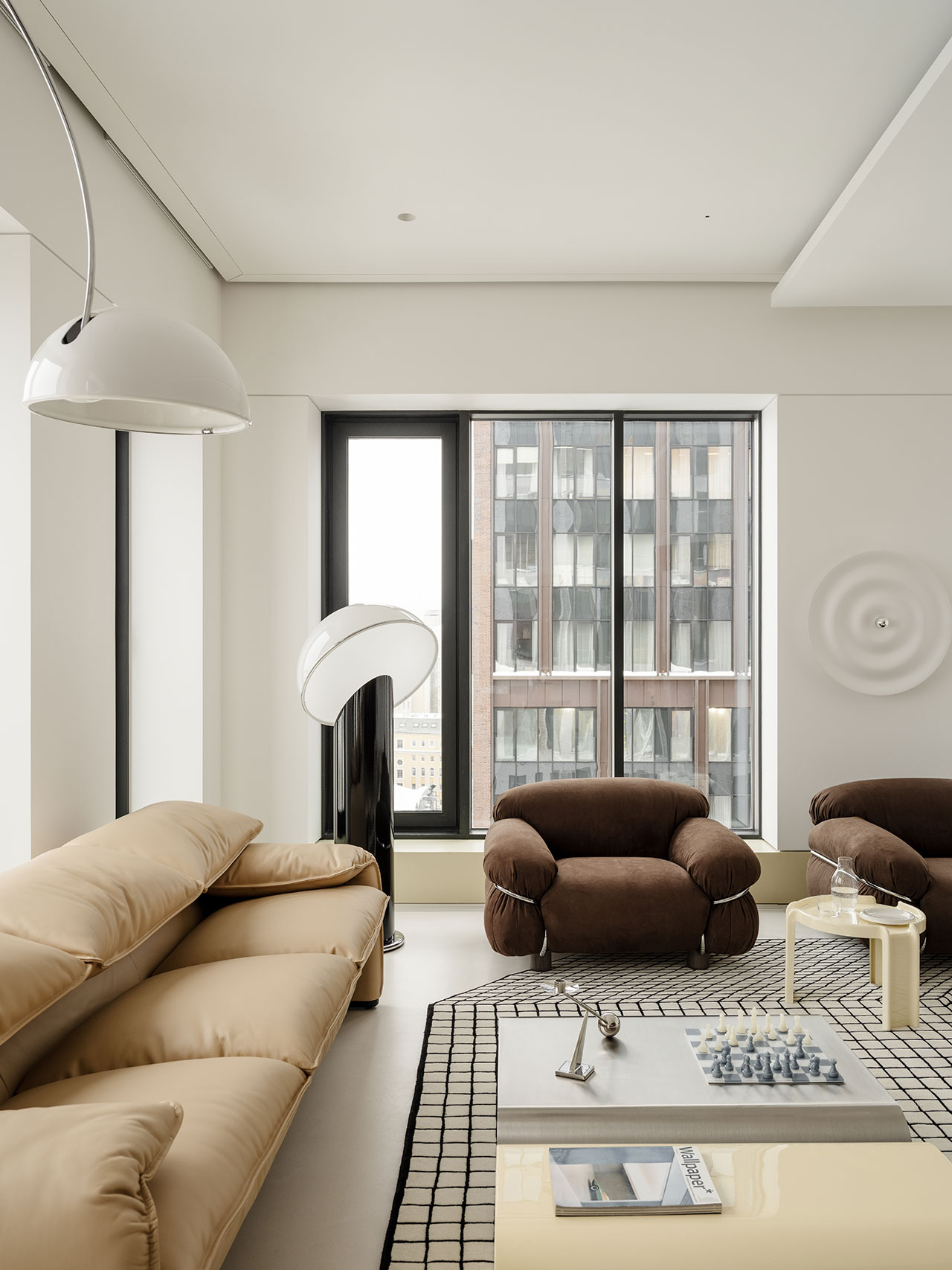 Modern Antwe living room with beige sofa, brown armchair, contemporary lamps, checkered rug, low tables, chess set, and large window overlooking city buildings.