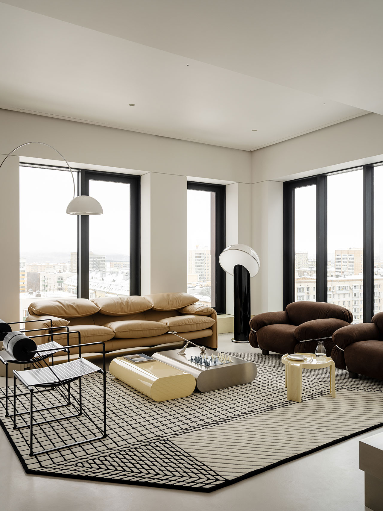 Modern living room with a tan sofa, two brown chairs, sculptural coffee tables, geometric rug, and large windows overlooking a stunning Antwe cityscape.