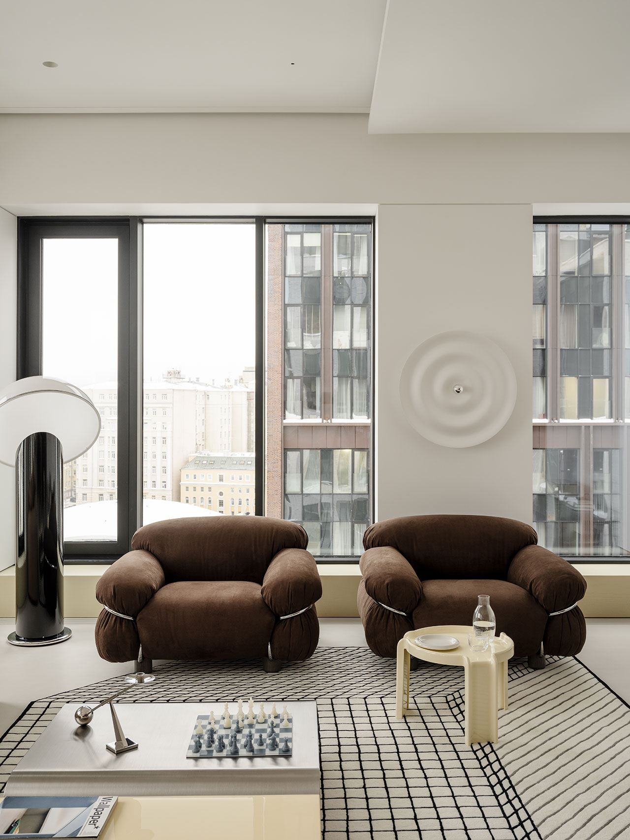 Modern Antwe living room with two brown armchairs, a small beige side table, a chess set on the coffee table, patterned rug, large windows, and minimalist decor.