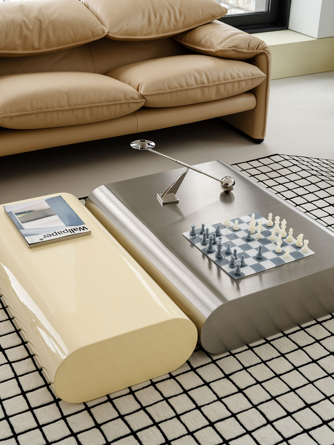 Modern Antwe living room with a beige sofa, grid-pattern rug, two minimalist tables—one cream, one metallic—holding a chessboard, a magazine, and a sleek silver lamp.