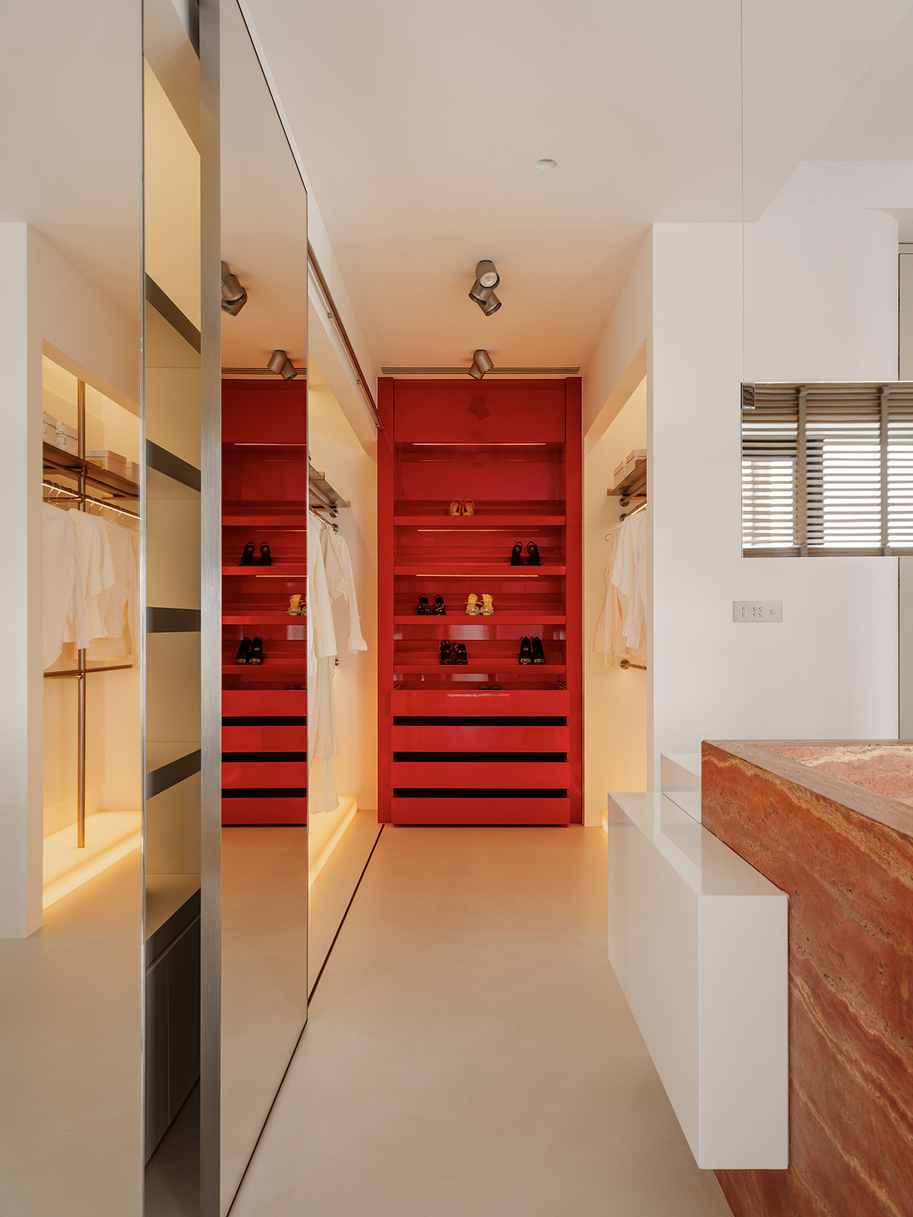 Modern Antwe walk-in closet with white walls, a red shelving unit displaying shoes and accessories, clothing hanging on both sides, and warm, recessed lighting.