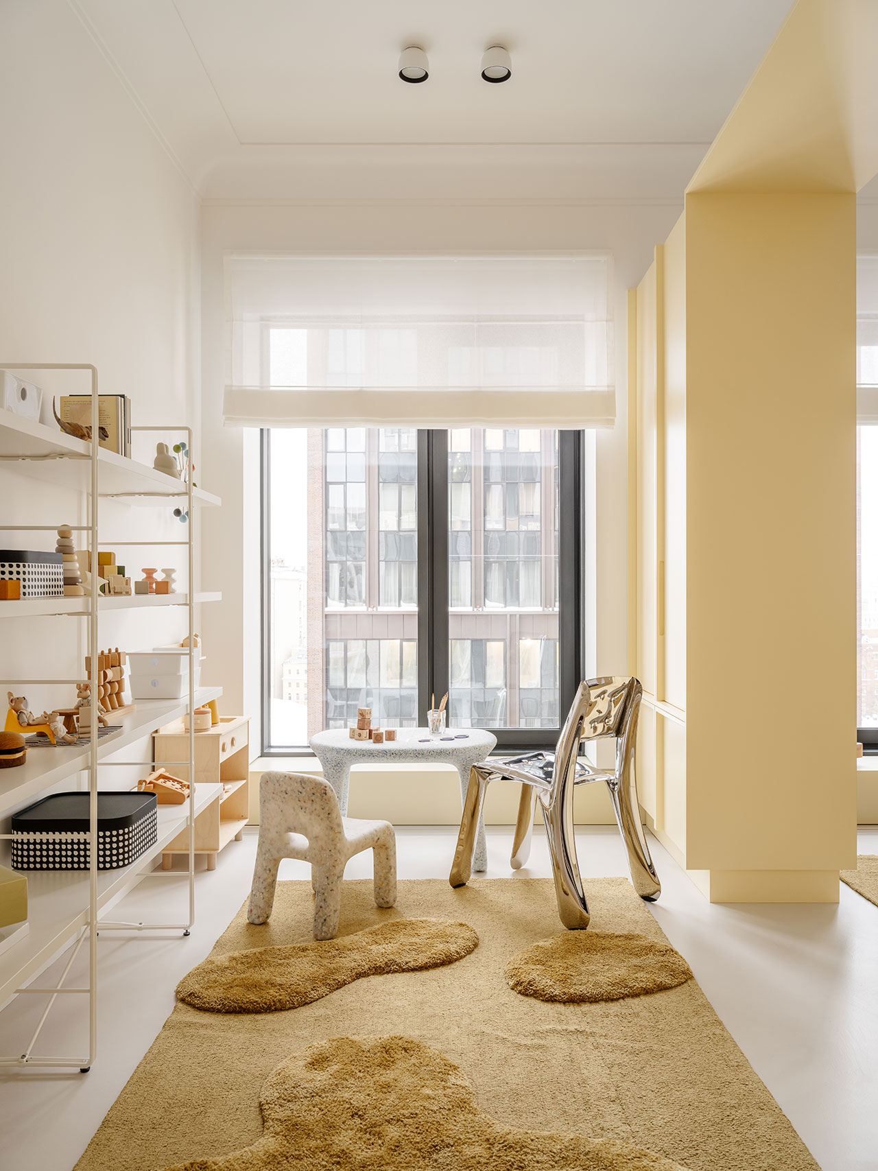 A modern children's room with soft yellow walls, abstract rugs, playful chairs, a small table by the window, and minimalist Antwe shelving adorned with toys and decor.