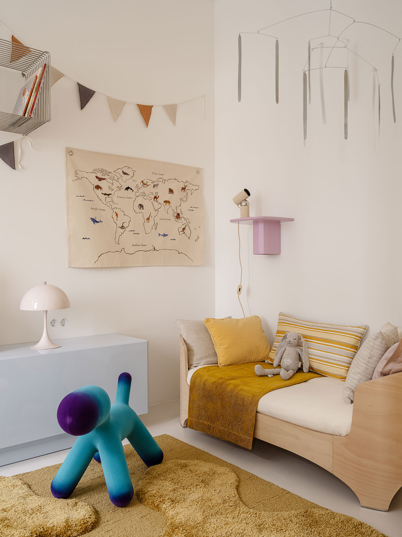 A cheerful children’s room in Antwe with a small sofa, stuffed toys, yellow blanket, wall map, triangular bunting, a blue animal-shaped chair, and a modern white lamp on a cabinet.