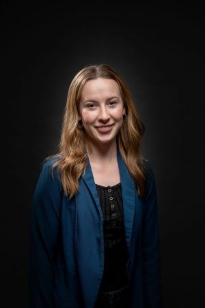 Sara Jobson wears a black shirt and blue blazer and stands in front of a black background.