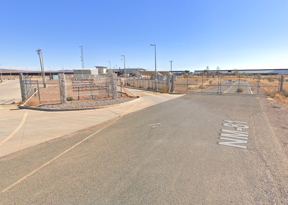 Exterior view of the Antelope Wells Border Station in New Mexico, featuring a gated entrance with a sign for U.S. Customs and Border Protection. The surrounding area is arid with sparse vegetation, and the sky is clear.