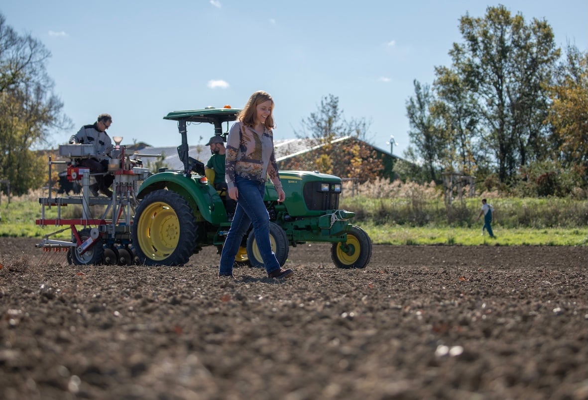A woman walks across a plowed field in front of a green tractor with two people operating machinery. Trees, a building, and another person are visible in the background under a blue sky.