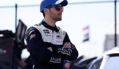 Racecar driver wearing a blue cap and sunglasses stands with arms crossed at a track behind a chain-link fence.