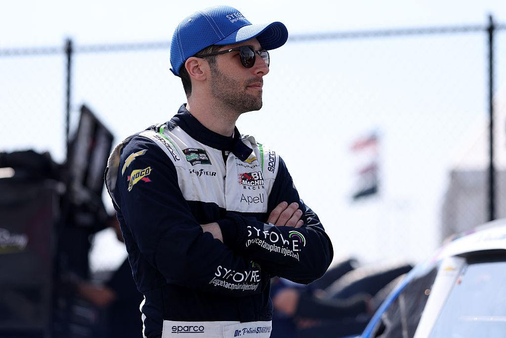 Racecar driver wearing a blue cap and sunglasses stands with arms crossed at a track behind a chain-link fence.