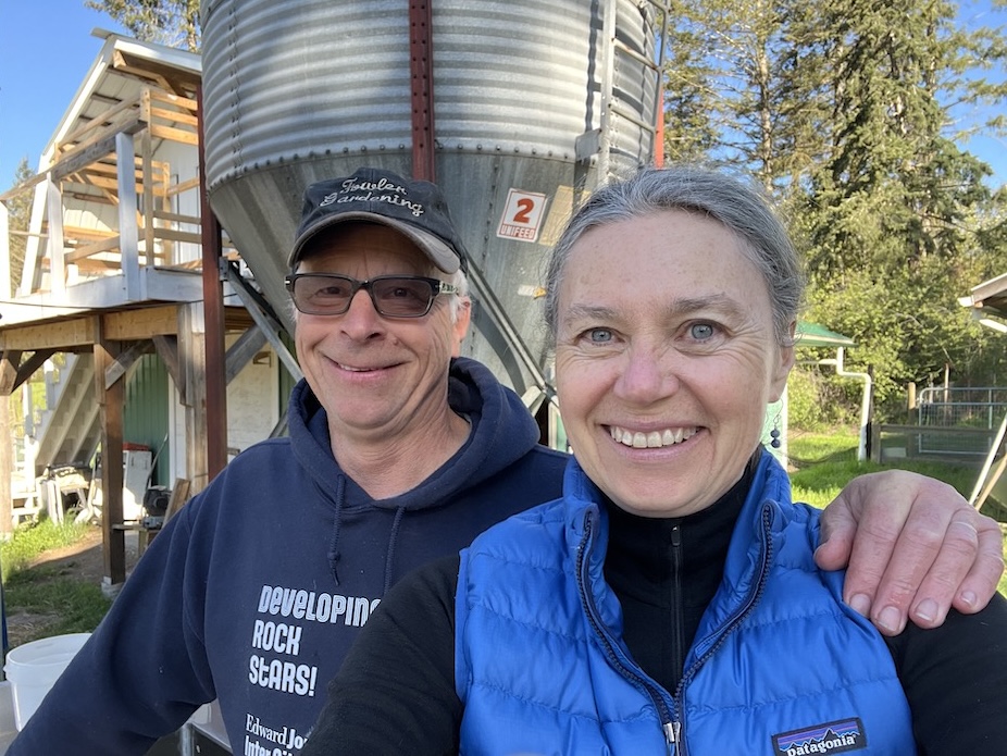A man and a woman sit in front of a large grey metal water tank on their property.