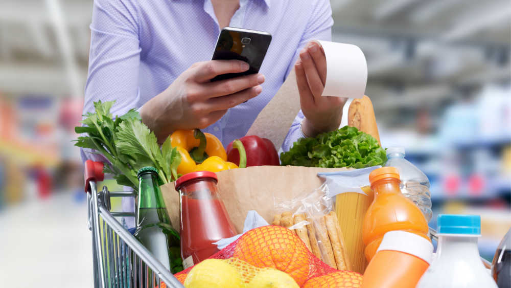 Person pushing a supermarket car full of groceries and looking at receipt and phone