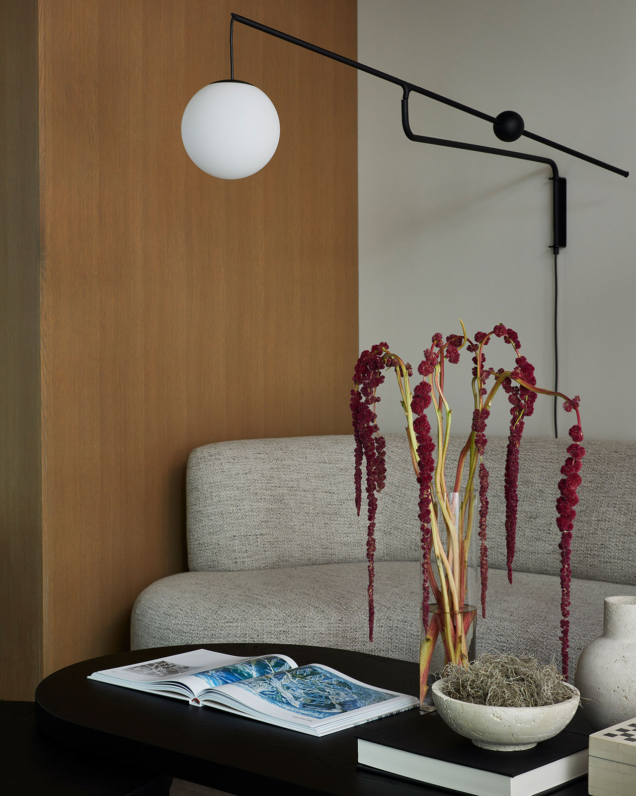 Modern living room with a gray sofa, black wall-mounted lamp, a vase of red amaranthus flowers, and an open book on a black coffee table.