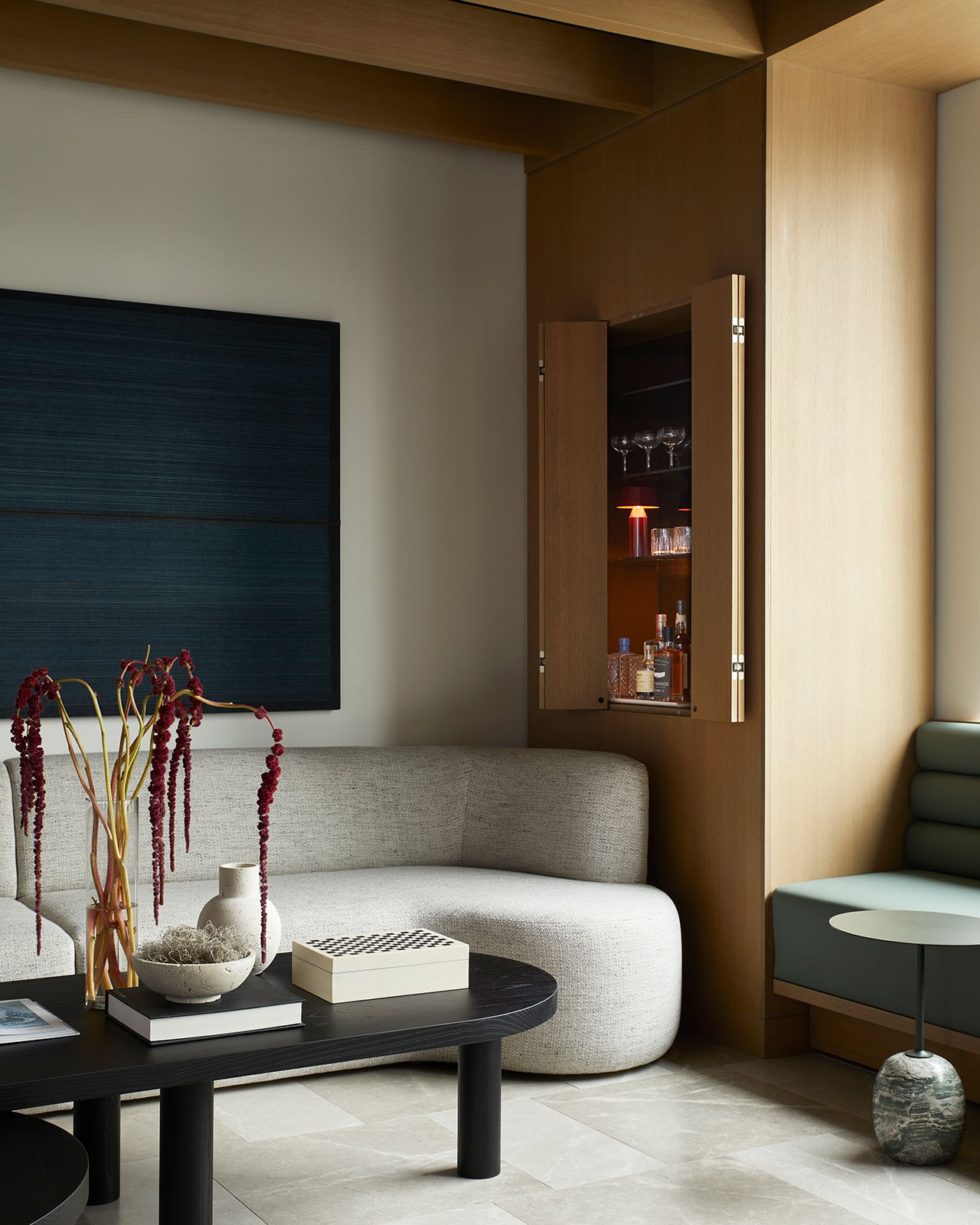 A modern living room with a curved sofa, a black coffee table holding books and decor, a wall bar with glassware, and a large blue artwork on the wall.