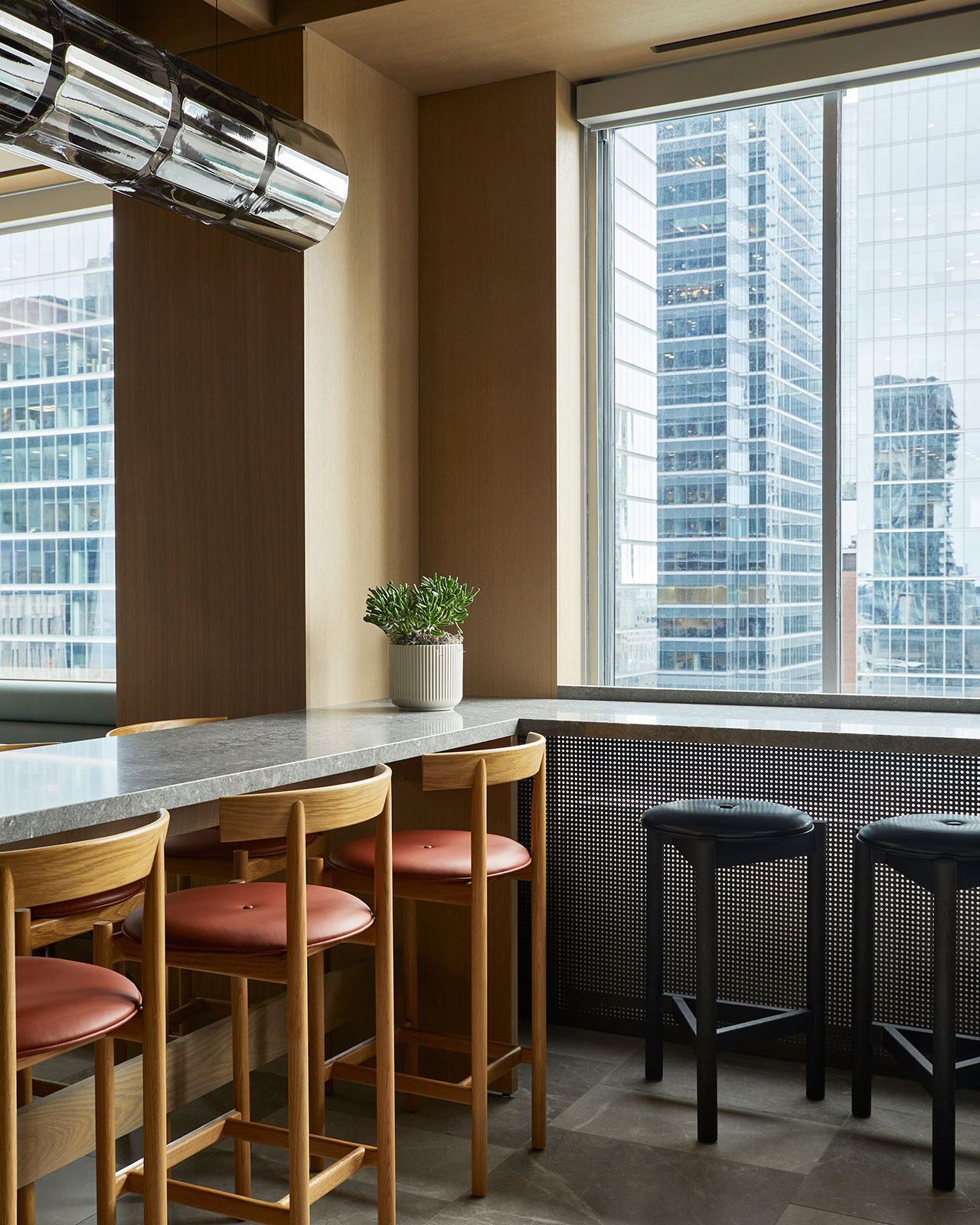 A modern dining area with wooden chairs, black stools, a marble countertop, a potted plant, and large windows overlooking tall city buildings.