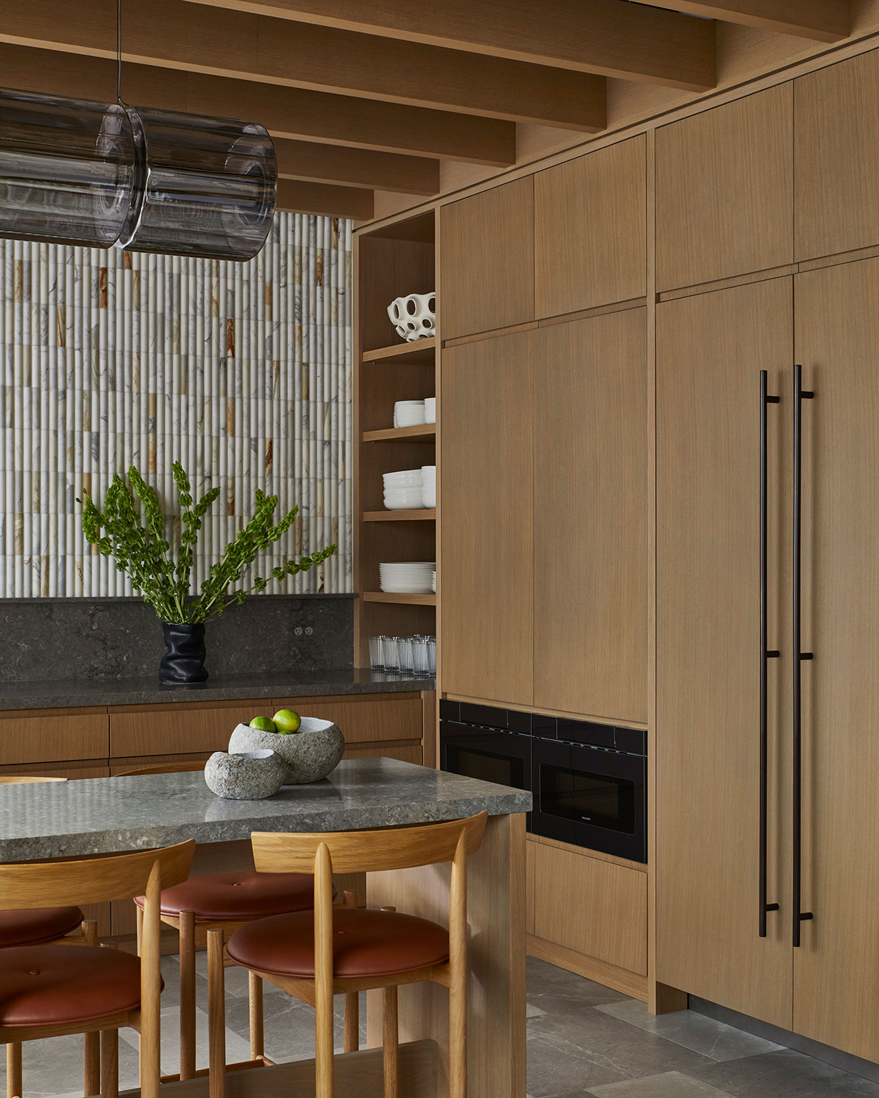 Modern kitchen with light wood cabinetry, open shelves with dishes, stone countertops, a black vase with greenery, and a bowl of apples on the island.